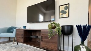 A living room with a flat-screen TV mounted on the wall, a wooden cabinet with a plant, a floor lamp, and a decorative vase.