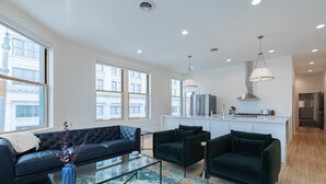 Ein modernes Wohnzimmer mit Sofa, zwei Sesseln, einem Couchtisch und einer Küchenzeile inklusive Kühlschrank und Herd.