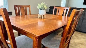 A wooden dining table with six chairs, a potted plant in the center, and a grey sofa in the background.