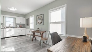 Een moderne keuken met witte kasten, een eethoek met een houten tafel en een groot raam met zonwering.