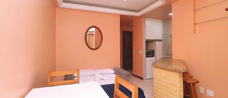 A hotel room with a bed, a wooden headboard, a small kitchen area, and a bathroom visible through an open door.