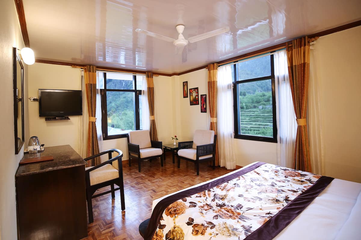 Habitación de hotel con cama, escritorio con televisor, dos sillas y ventanas amplias con cortinas.