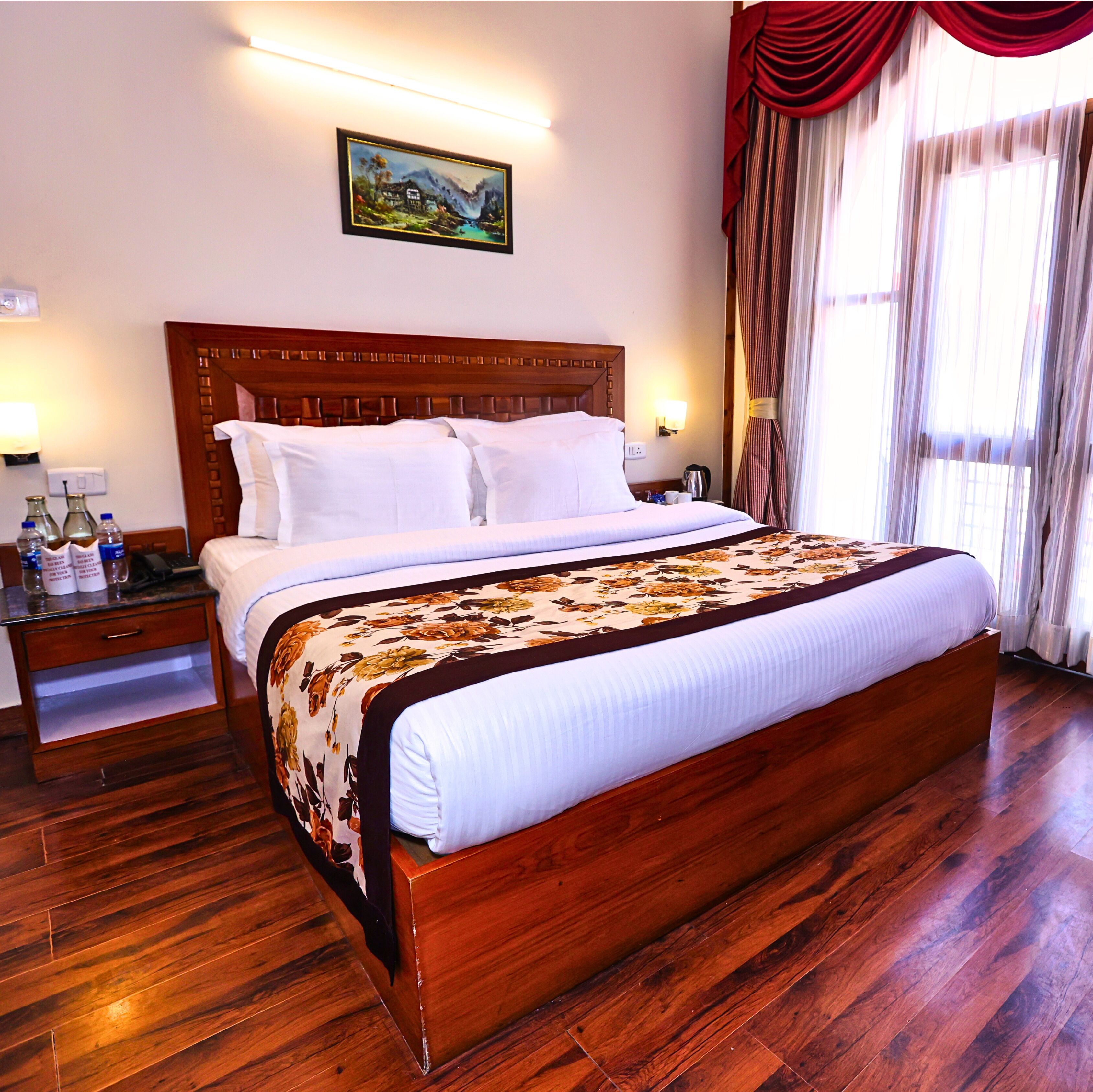 A hotel room with a wooden bed, white bedding, a floral bedspread, a nightstand with a lamp, and a painting on the wall.