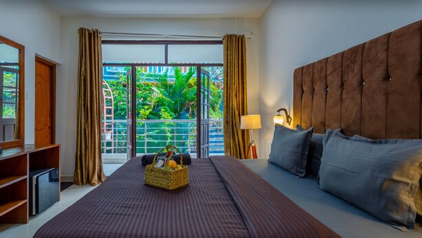 A hotel room with a large bed, a wooden headboard, a bedside table with a lamp, and a view of greenery through the window. - The Baga Village (Baga)