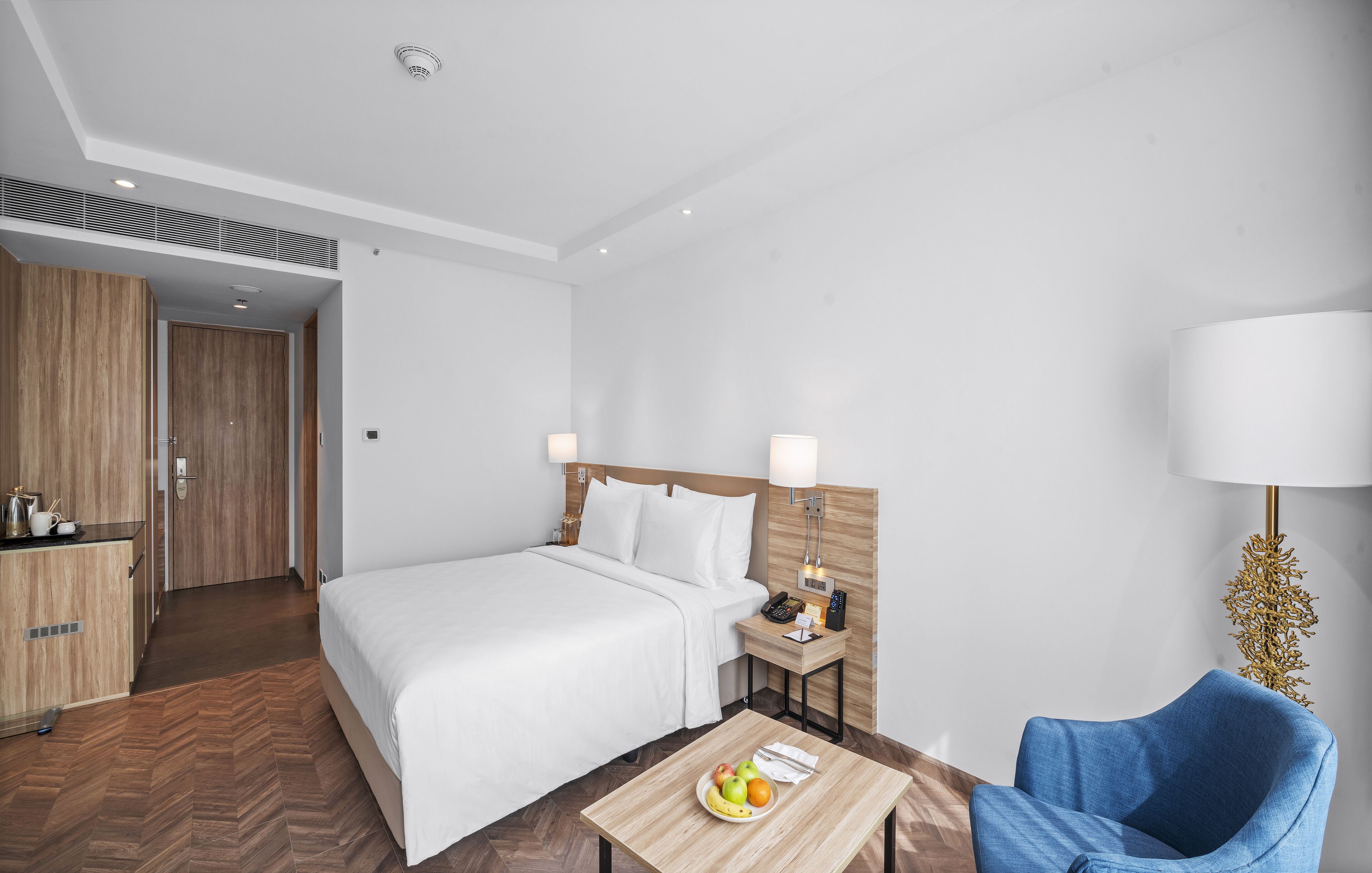 Une chambre d’hôtel avec un lit, une table de chevet, une lampe, une chaise et une petite table sur laquelle se trouvent des fruits.