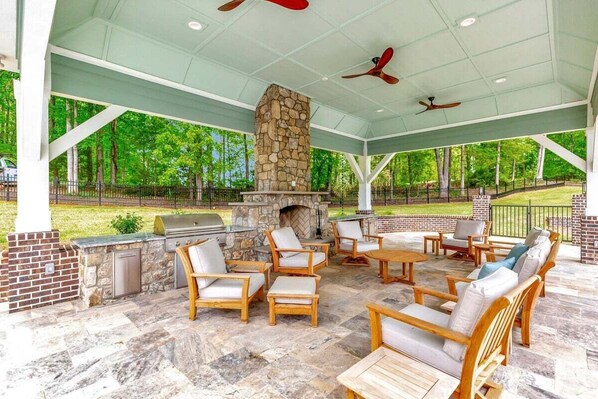An outdoor patio with stone flooring, wooden furniture, a stone fireplace, and ceiling fans.
