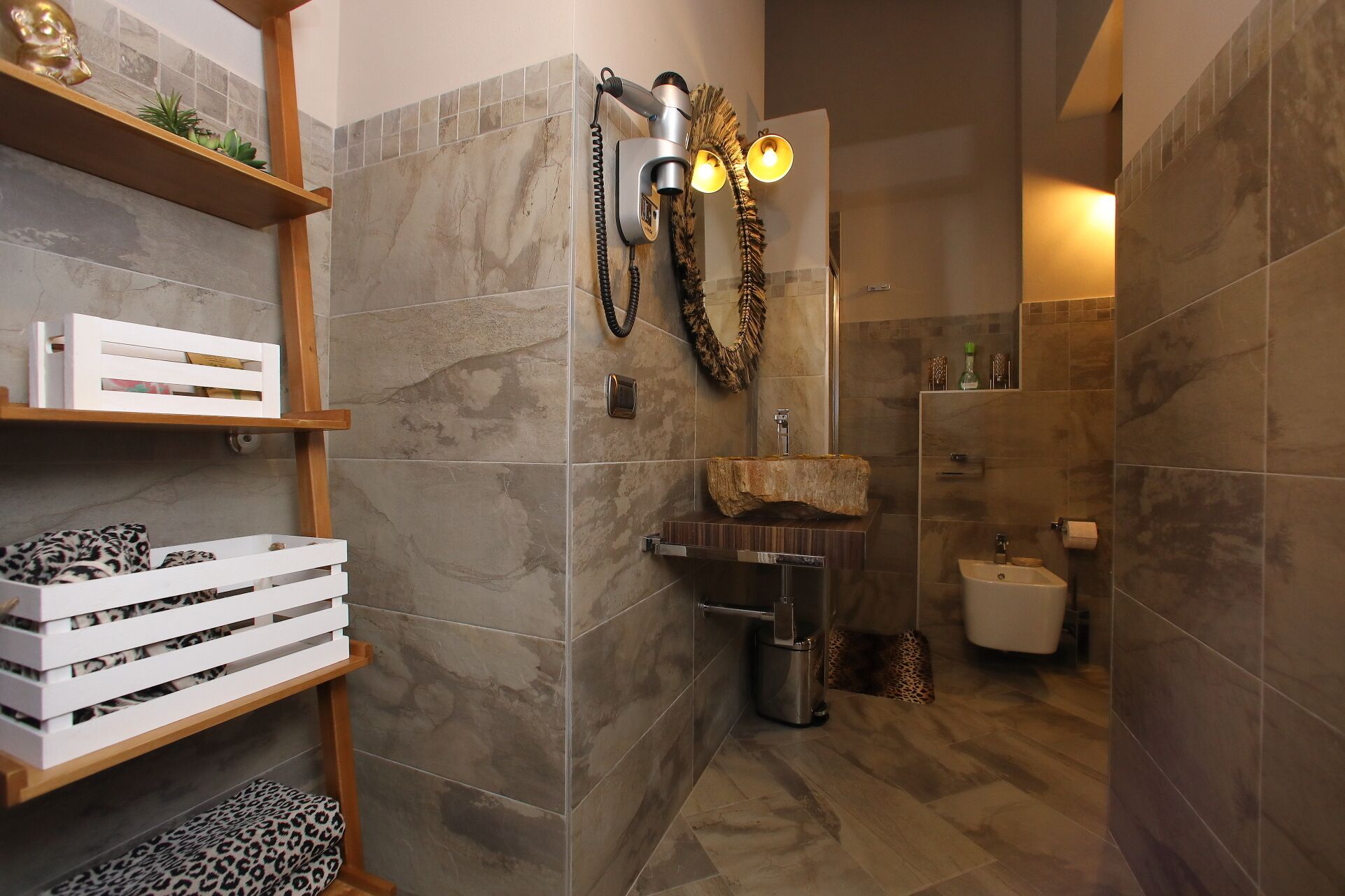 Une salle de bain moderne avec des carreaux de marbre, une étagère en bois avec des boîtes, un lavabo original et une douche équipée d’un dispositif fixé au mur.