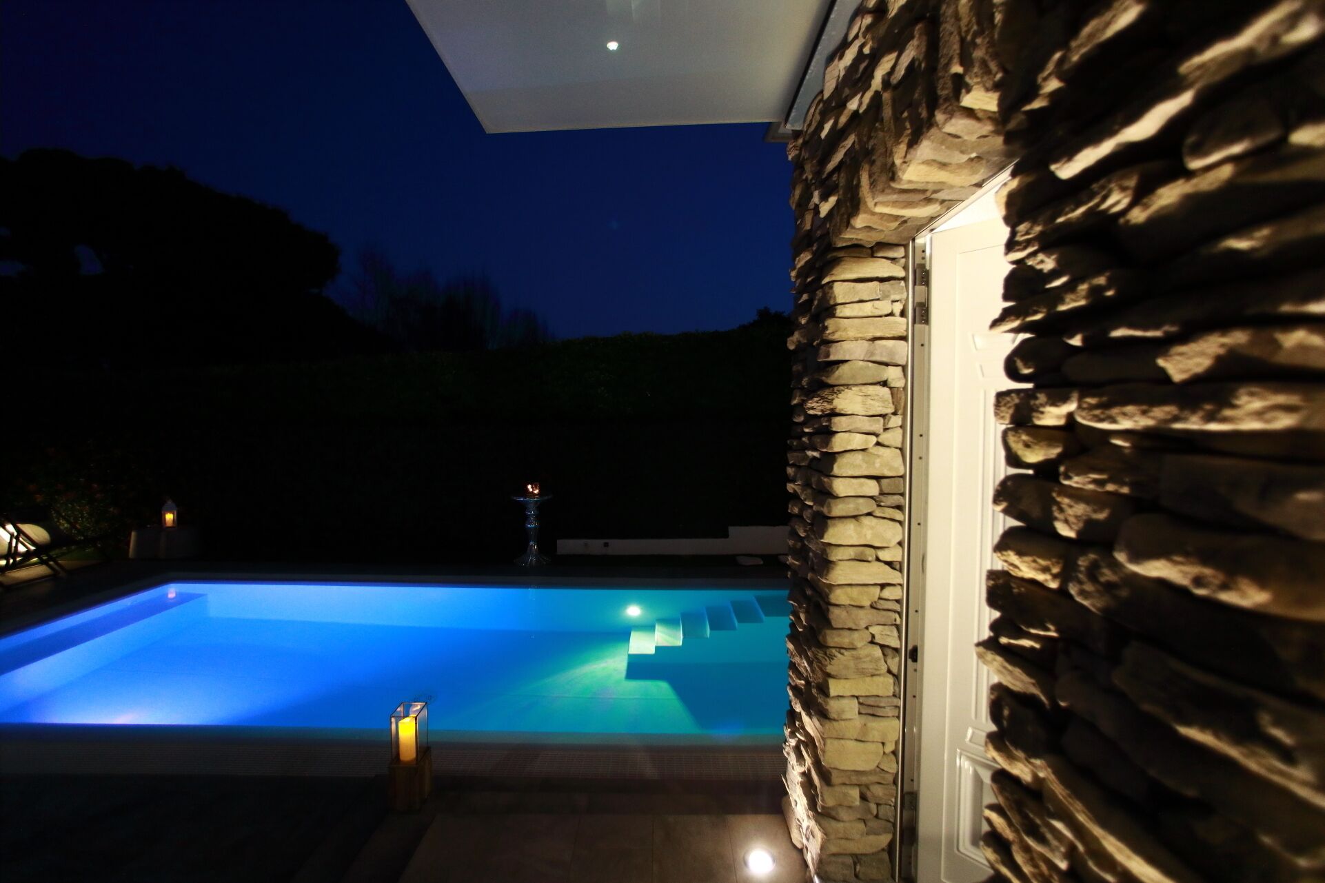 A swimming pool with a stone wall and a glass door, surrounded by a dark night sky.