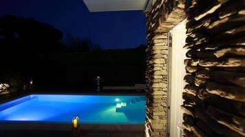A swimming pool with a stone wall and a glass door, surrounded by a dark night sky.