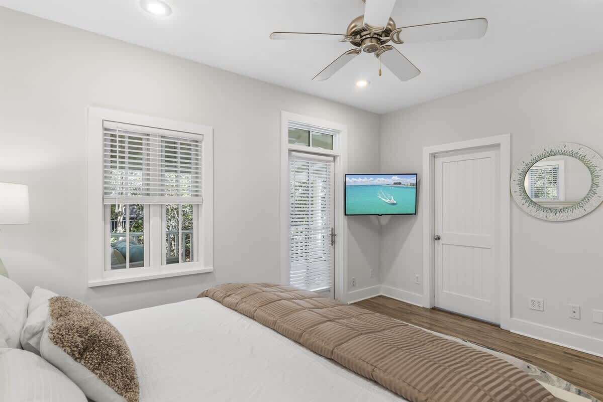 A bedroom with a bed, a television, a ceiling fan, and two windows with blinds.