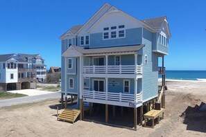 Una casa fronte mare a due piani con un balcone, un pontile di legno e una spiaggia sabbiosa sullo sfondo.