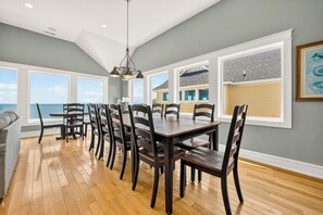 A dining area with a view of the ocean, wooden flooring, and a large window with blinds.