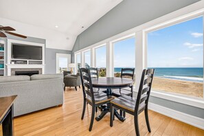 A room with a dining table and chairs, a sofa, a television, and a fireplace. Large windows offer a view of the beach and ocean. - Cape Point, charming 7-Bedroom Oceanfront + Pets Stay Free! (Buxton)