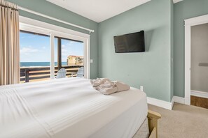 A hotel room with a large bed, a TV mounted on the wall, and a view of the beach through the window.