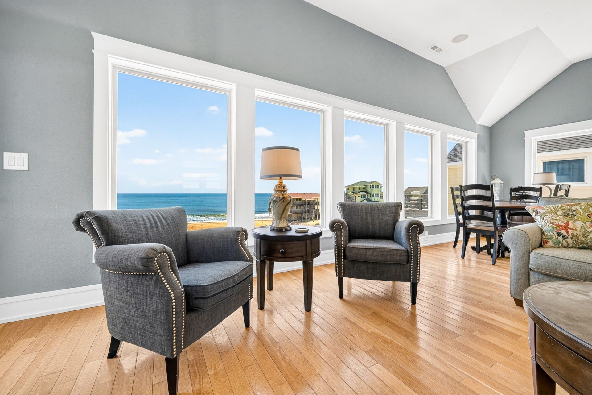 A spacious living room with a high ceiling, large windows offering a view of the ocean, and a dining area with wooden flooring.