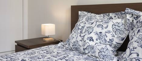 A neatly made bed with a floral-patterned bedspread and matching pillows, a bedside lamp, and a wooden nightstand.