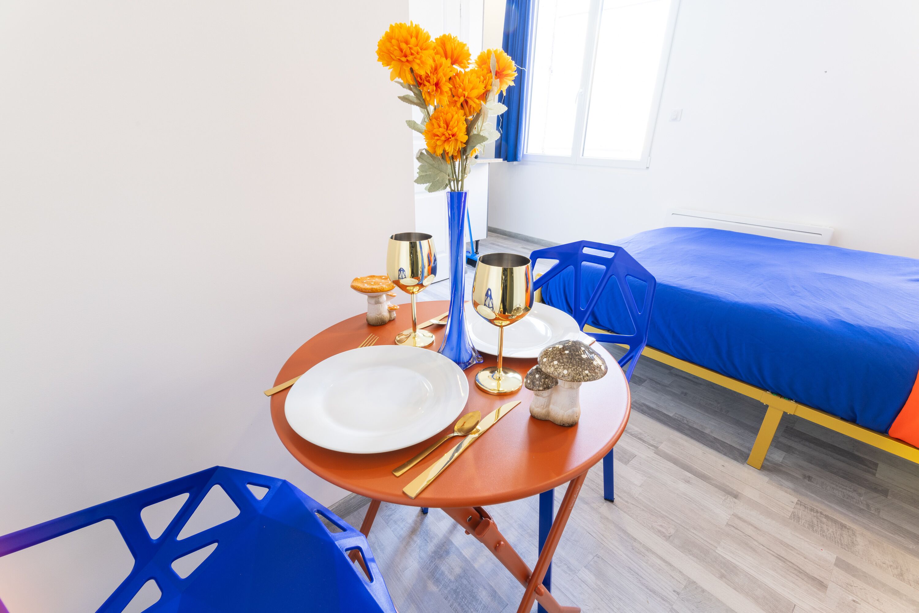 Une table à manger bien mise, avec une assiette blanche, des couverts dorés, un verre et un vase de fleurs. On aperçoit, à l’arrière-plan, un lit recouvert d’une couverture bleue.