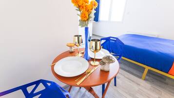 Une table à manger bien mise, avec une assiette blanche, des couverts dorés, un verre et un vase de fleurs. On aperçoit, à l’arrière-plan, un lit recouvert d’une couverture bleue.