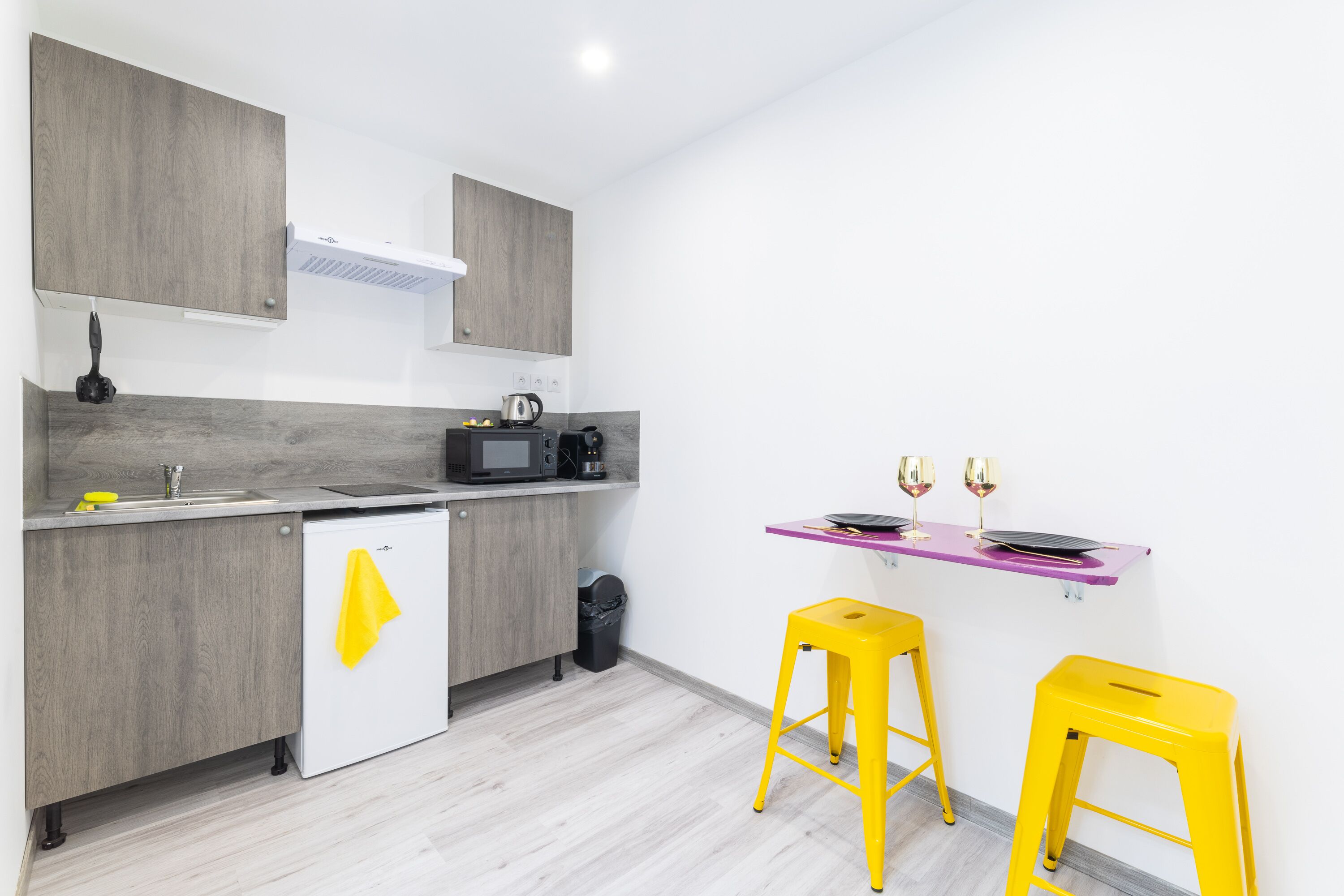 A modern kitchen with grey cabinets, a white refrigerator, a microwave, and a coffee maker. There are two yellow stools and a purple shelf with glasses.