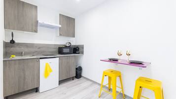 A modern kitchen with grey cabinets, a white refrigerator, a microwave, and a coffee maker. There are two yellow stools and a purple shelf with glasses.
