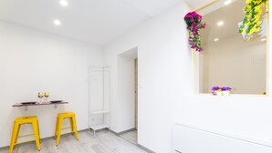 A modern room with white walls, a wooden floor, two yellow stools, a small table with a wine glass, and a mirror with flowers. - GLM Residences 2 (Saint-Quentin)