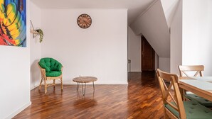 A room with a wooden floor, a green chair, a small round table, and a wooden dining table with chairs. There is a wall clock and a colorful painting. - RentPlanet - Apartament Odrzańska (Wroclaw)