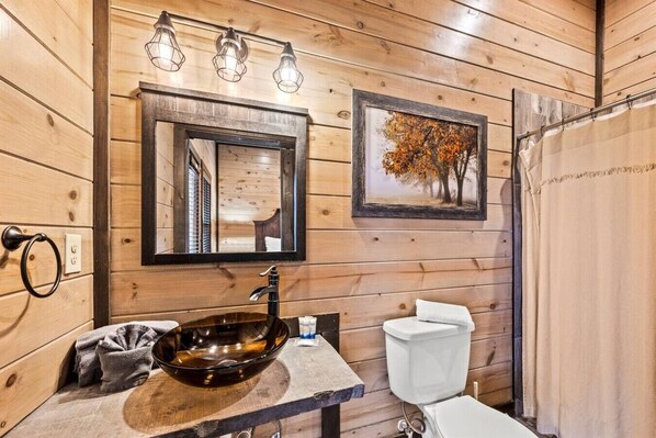 A wooden cabin bathroom with a large mirror, a vessel sink, a toilet, and a framed picture of trees. - 8 Bedroom ~Mtn View~ Sleeps 22 ~ Private Pool! (Sevierville)