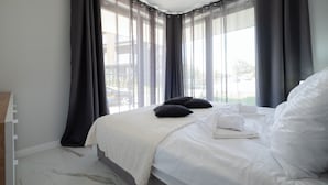 A neatly made bed with white linens and two black pillows, a white throw blanket, and a view of a balcony through sheer curtains. - Apartamenty Sun & Snow Żeglarska (gmina Lodygowice)