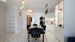 A modern kitchen with white cabinets, a black countertop, and stainless steel appliances. There is a dining area with a marble floor and black chairs. - Apartamenty Sun & Snow Żeglarska (gmina Lodygowice)