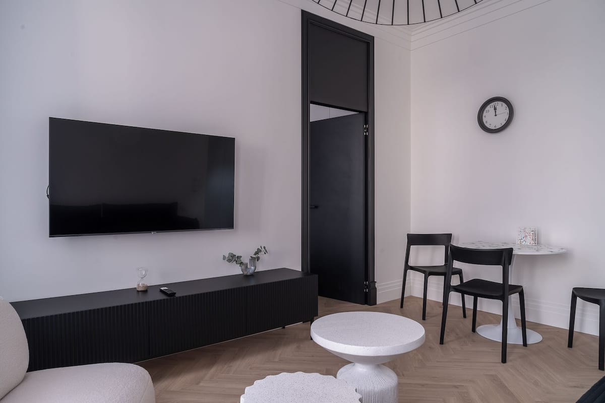 A modern living room with a flat-screen TV, a dark wooden console, a round white table, and two black chairs.