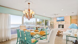 A dining area with a table set for four, surrounded by teal chairs, a chandelier, and a view of the water. - Ground level bay-front home on deep waterfront!  (Dauphin Island)