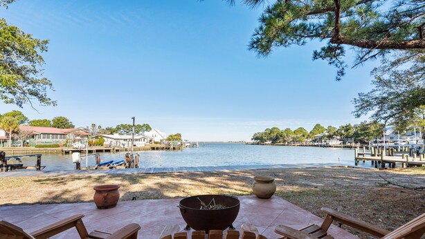 A lakeside patio with wooden chairs and a fire pit, overlooking a calm lake and dock area. - Ground level bay-front home on deep waterfront!  (Dauphin Island)