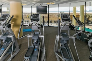 Ein Fitnessstudio mit Laufbändern und Trainingsgeräten, großen Fenstern mit Blick auf den Strand und einem Fernsehbildschirm.
