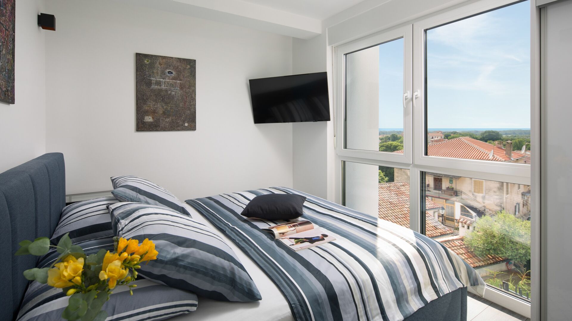 Ein Schlafzimmer mit Bett, Flachbildfernseher, Fenster mit Stadtblick und einem Bild an der Wand.