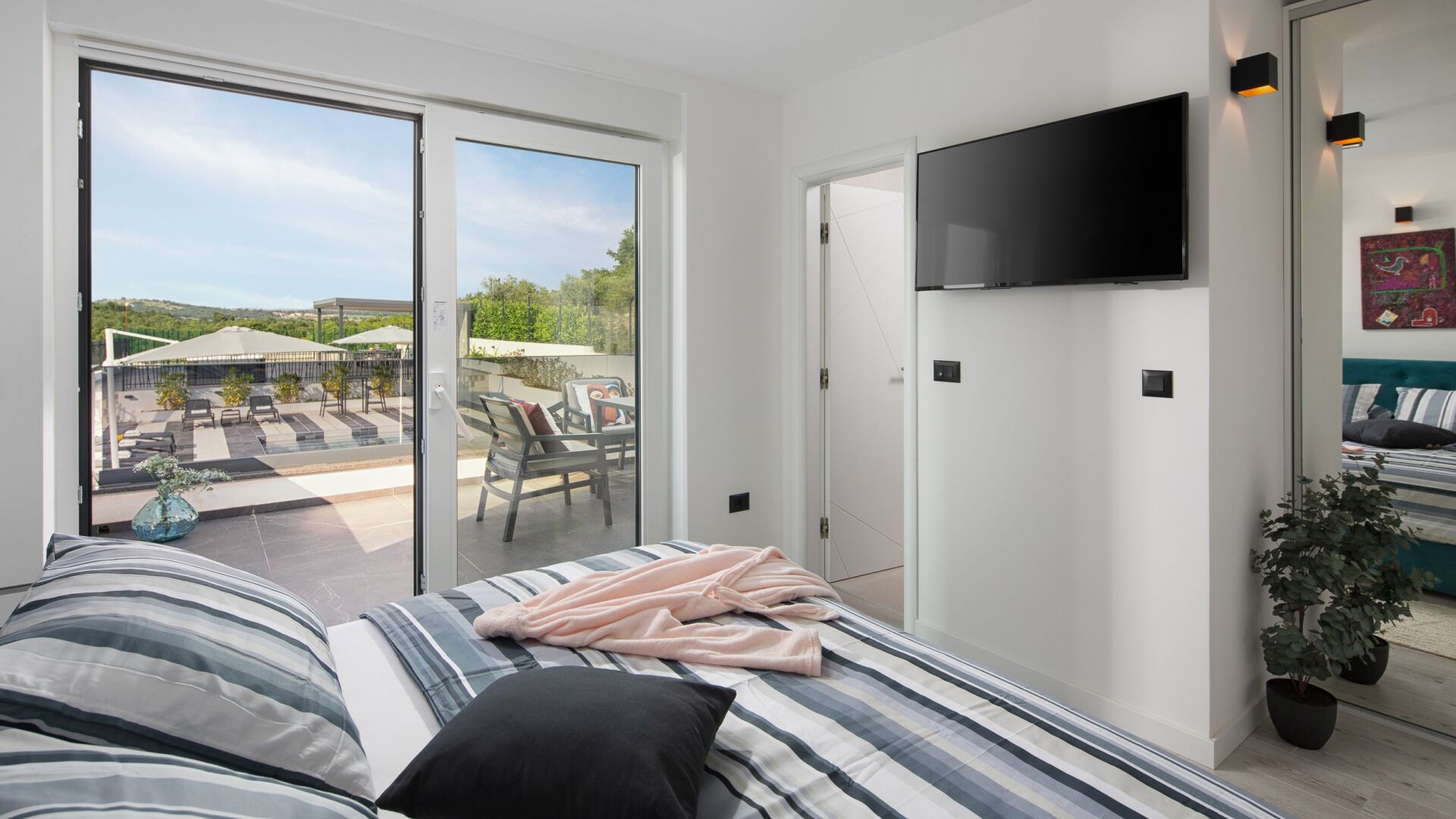 A bedroom with a large bed, a TV mounted on the wall, and a sliding glass door leading to an outdoor patio with chairs and umbrellas.