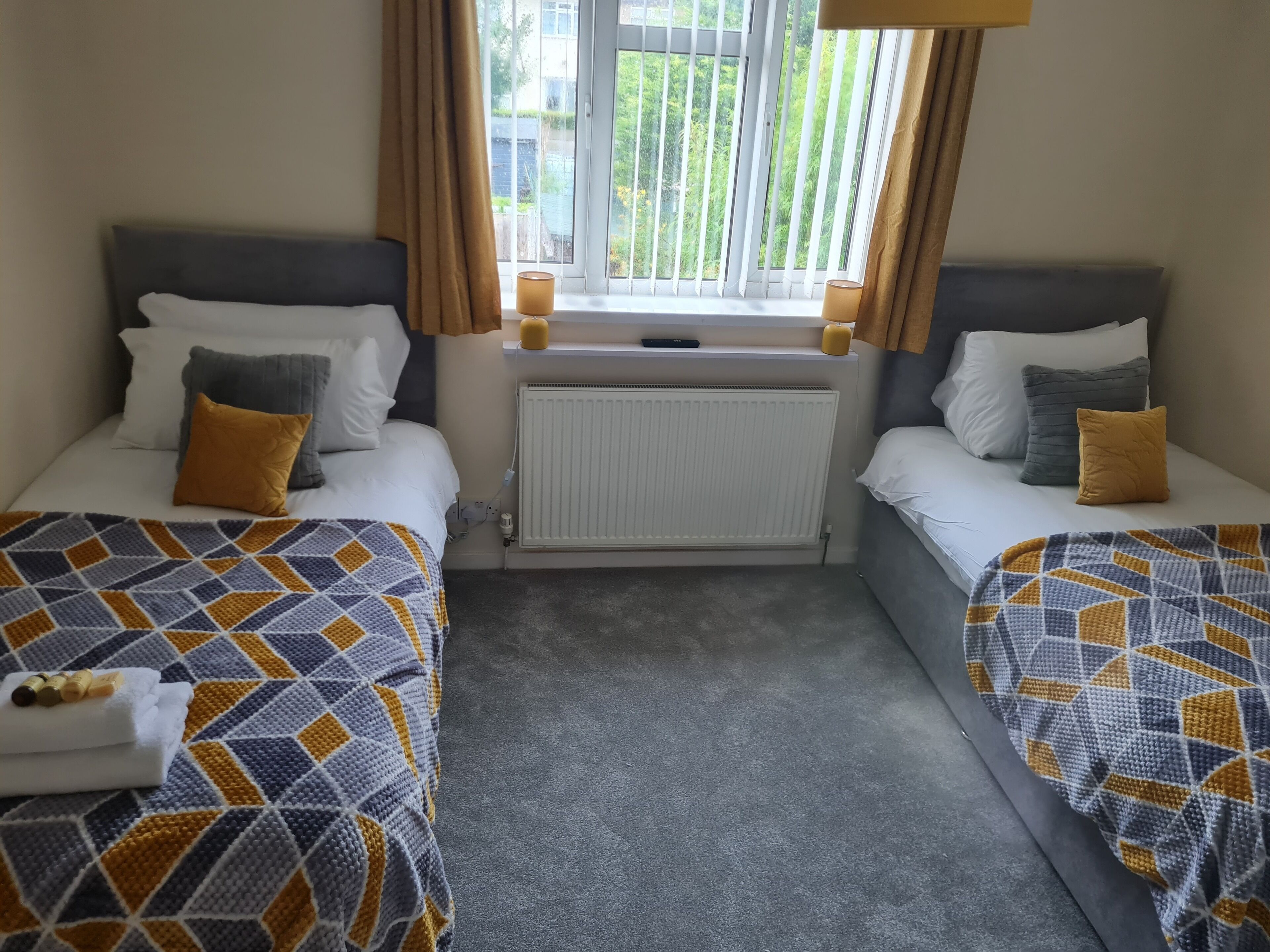 A hotel room with two beds, a window with curtains, a radiator, and a small table with a towel.