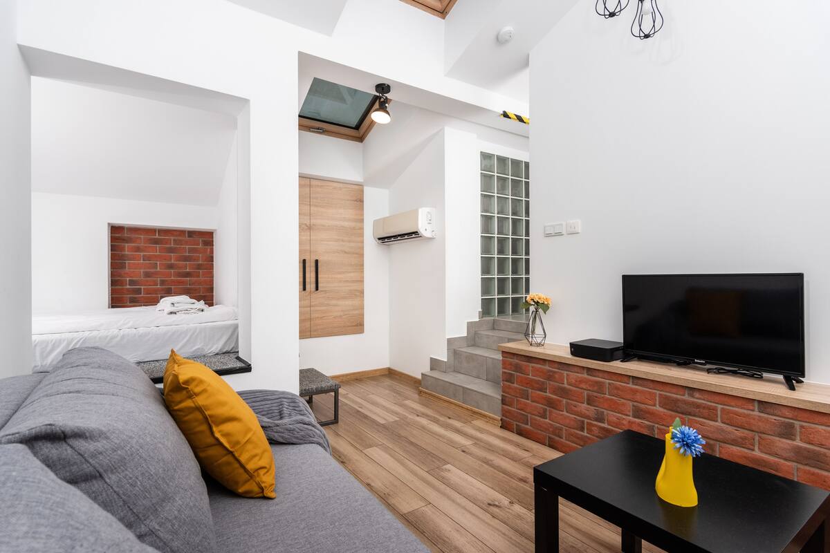 A modern living room with a grey sofa, a yellow pillow, a black coffee table with a yellow vase, a TV, and a brick fireplace.
