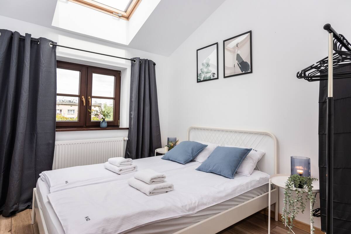 A bedroom with a bed, two pillows, a bedside table with a lamp, a window with curtains, and a view of buildings outside.