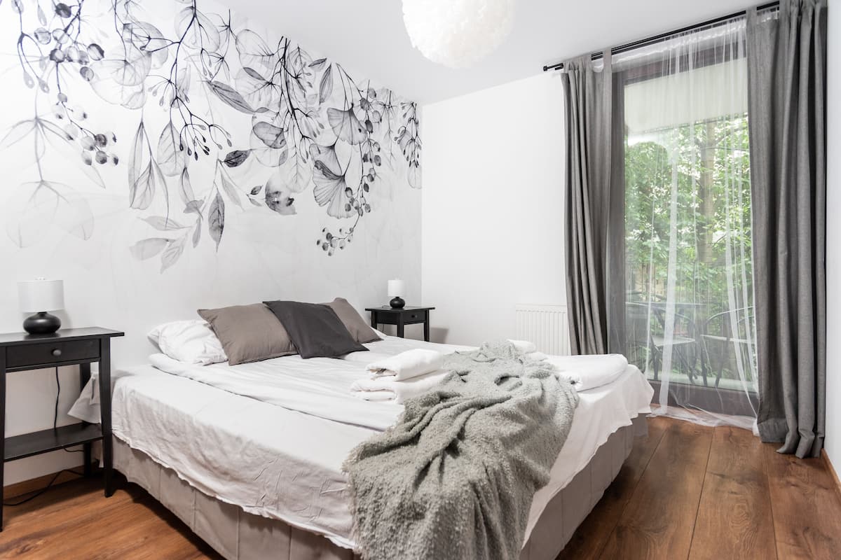 A bedroom with a bed, bedside tables, a wall mural, a window with curtains, and a view of greenery outside.