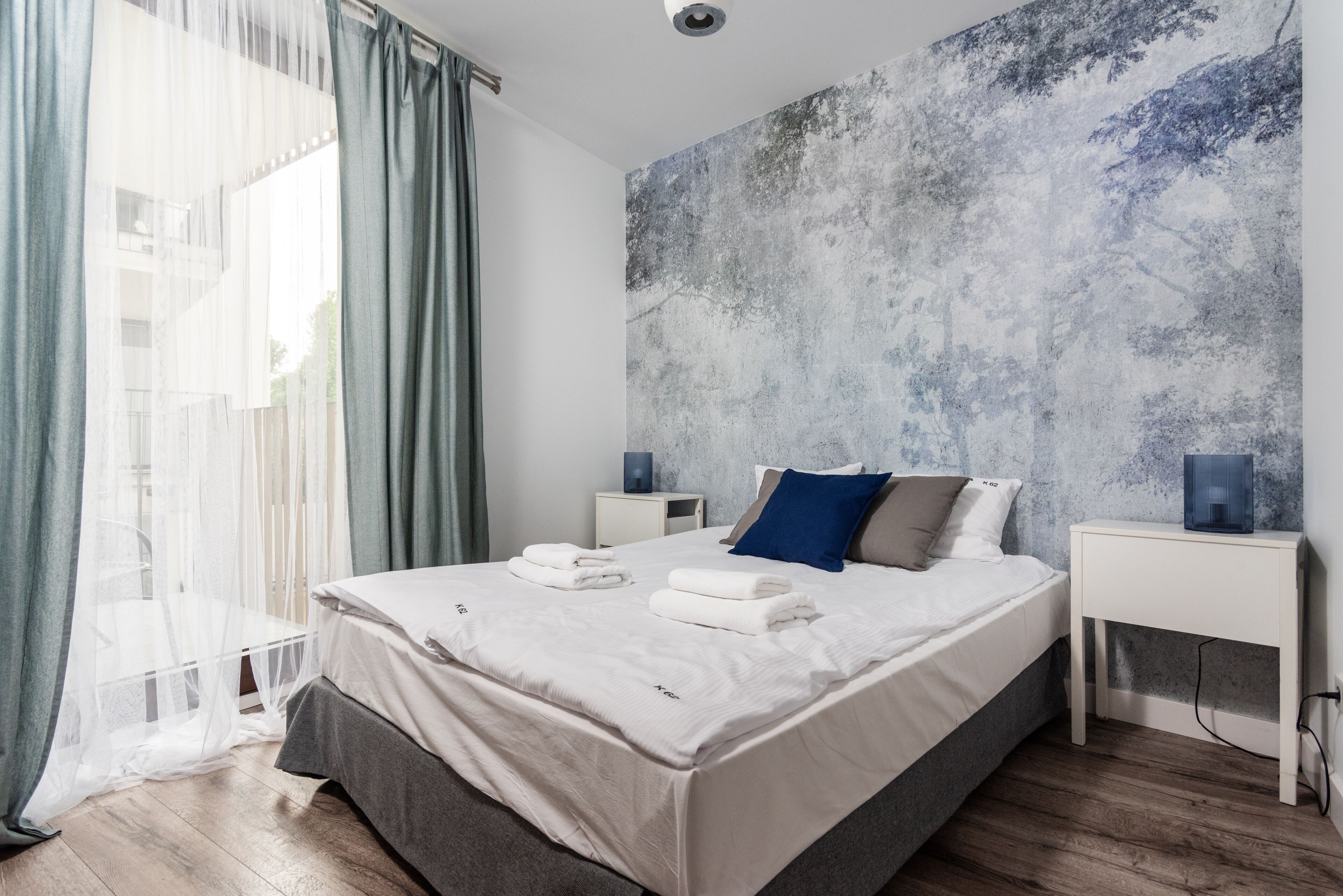 Une chambre moderne avec un grand lit, deux tables de chevet, une commode, une fenêtre avec des rideaux légers et un mur à texture.