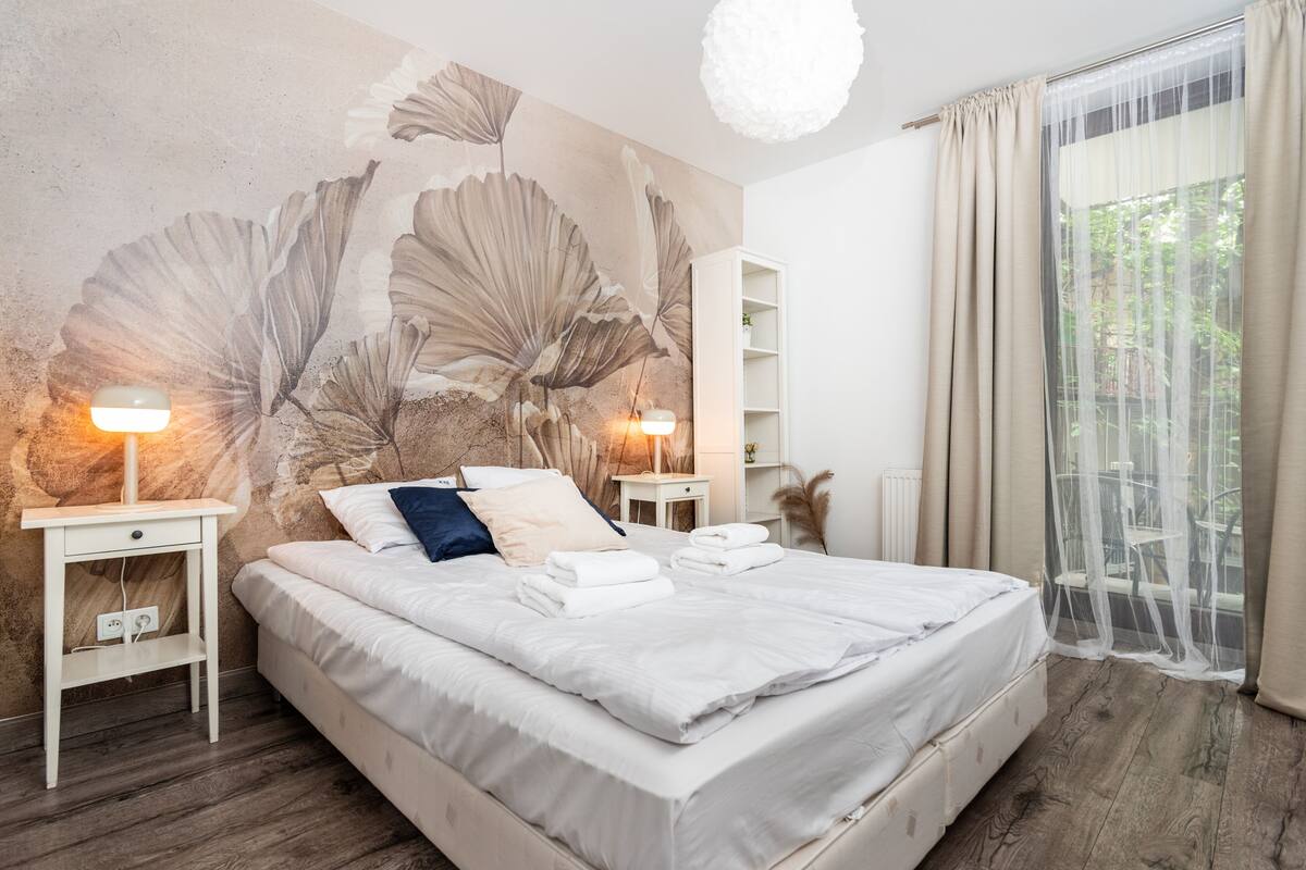 A bedroom with a large bed, two bedside tables, a wall mural, a window with curtains, and a hanging light fixture.