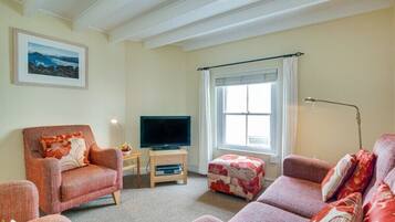 Una sala de estar con sofá, sillón y reposapiés, un televisor sobre un mueble bajo y una ventana con cortinas.