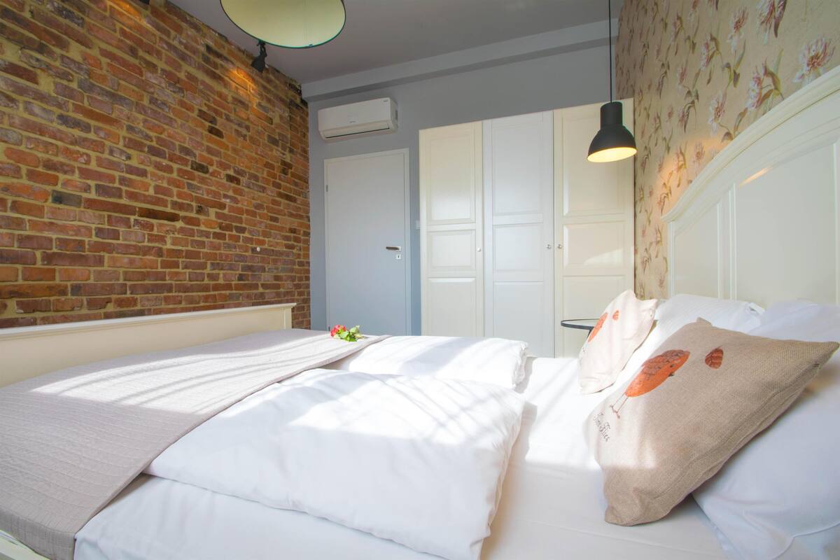 Une chambre avec un mur en briques, un lit recouvert de draps blancs, un luminaire suspendu et une porte donnant sur une salle de bain.