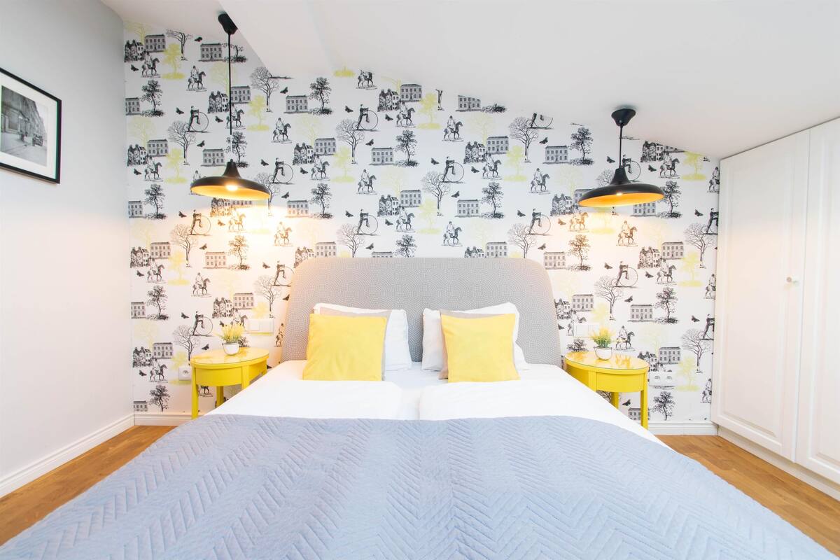 Une chambre à coucher comprenant un lit, deux tables de chevet, une petite table, une lampe fixée au mur et un papier peint à motifs.