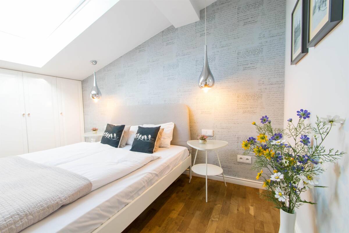 Une chambre moderne avec un grand lit, deux lampes suspendues, une petite table ronde et un mur orné d’un motif texturé.