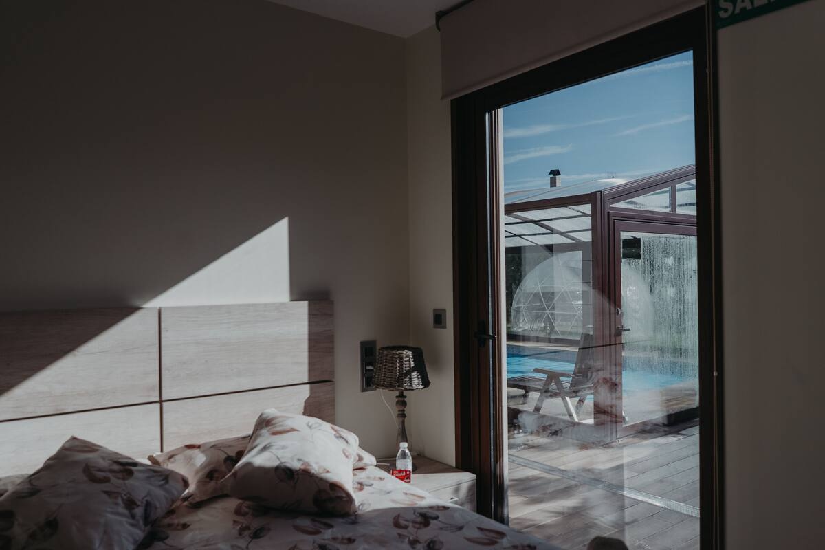 Une chambre moderne avec un grand lit, une table de chevet avec une lampe et une vue sur une piscine extérieure, visible à travers une porte vitrée.