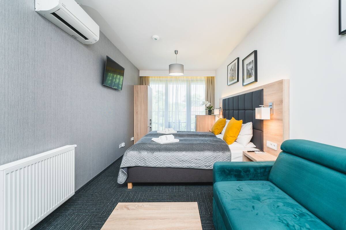 Quarto de hotel moderno com uma cama, um sofá verde-água, uma mesa de cabeceira de madeira e uma televisão fixada na parede.