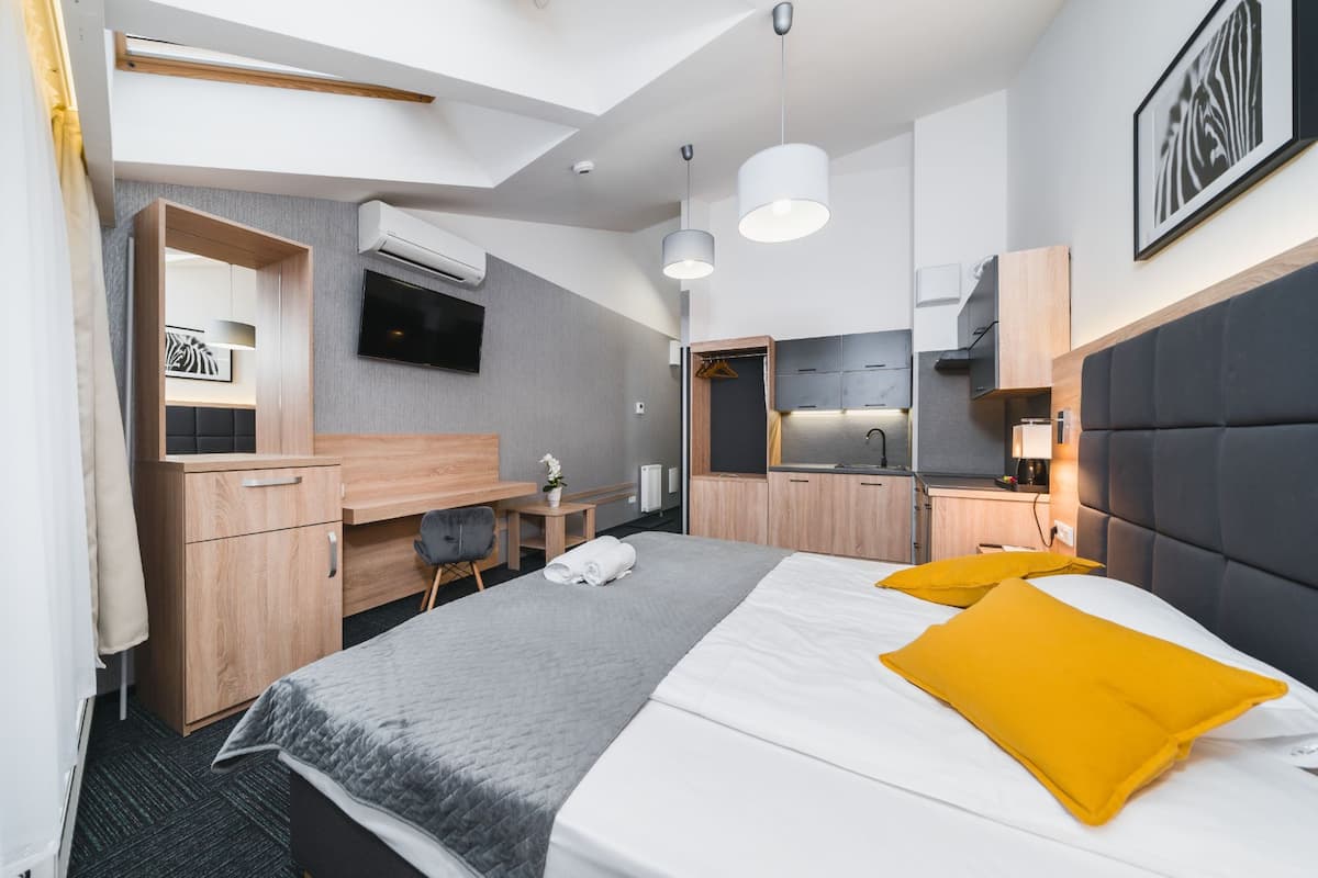 Quarto de hotel moderno com cama, escrivaninha, cadeira, televisão e cozinha compacta.