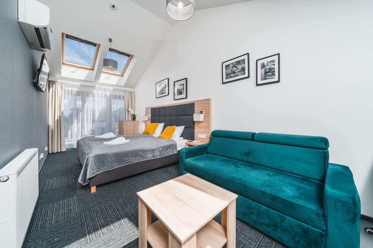 Quarto de hotel moderno com cama, sofá verde-água, mesa de madeira pequena e uma janela grande com cortinas.