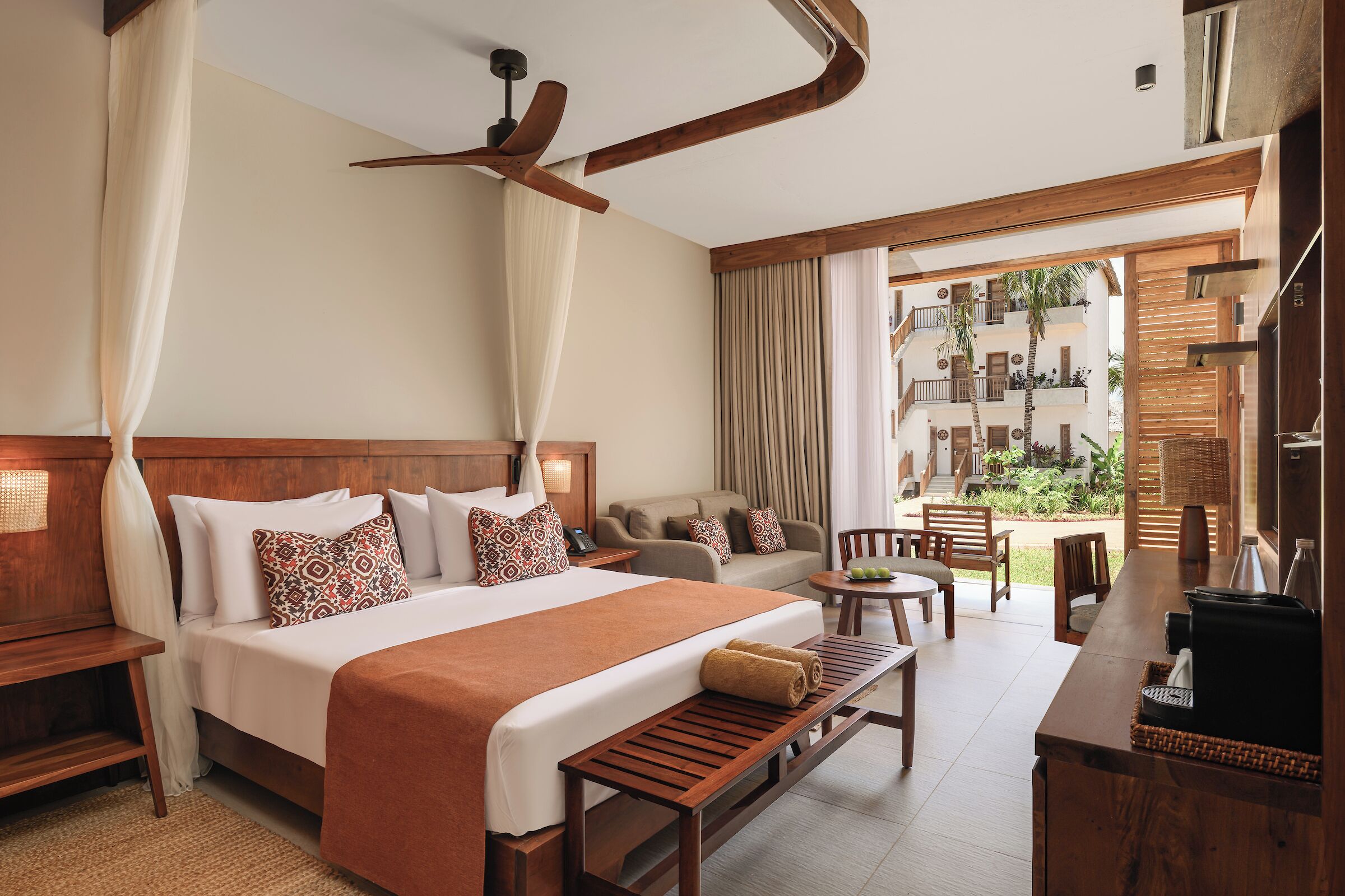 Habitación de hotel con una cama grande, un banco de madera, un escritorio y un balcón con vistas del edificio.